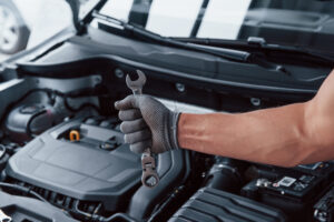 Man's hand in glove holds wrench in front of broken automobile.